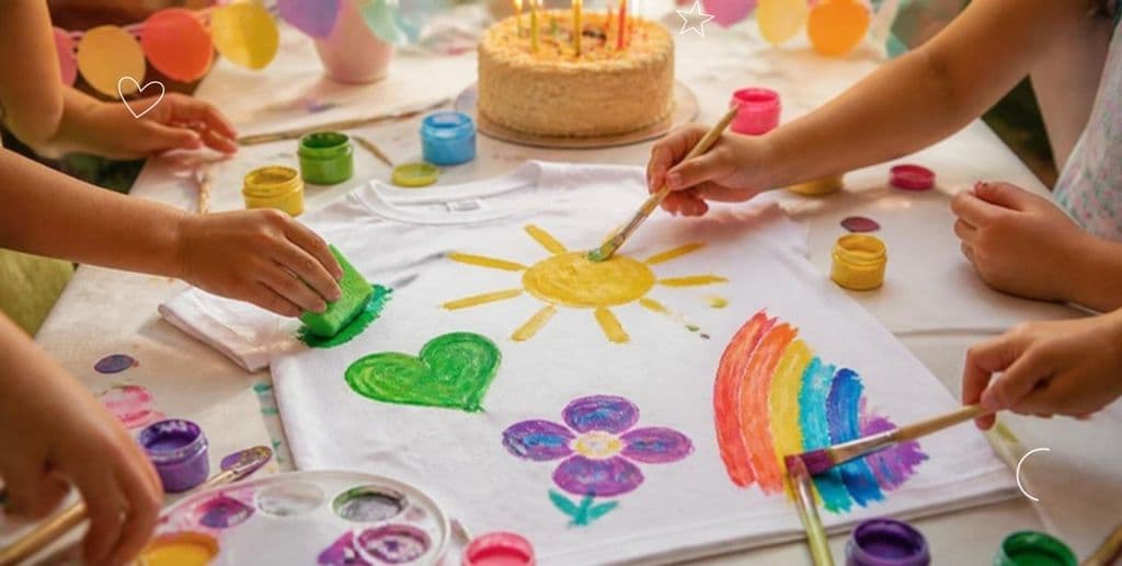 Mani di bambini che dipingono una maglietta bianca a una festa di compleanno, con la torta sullo sfondo